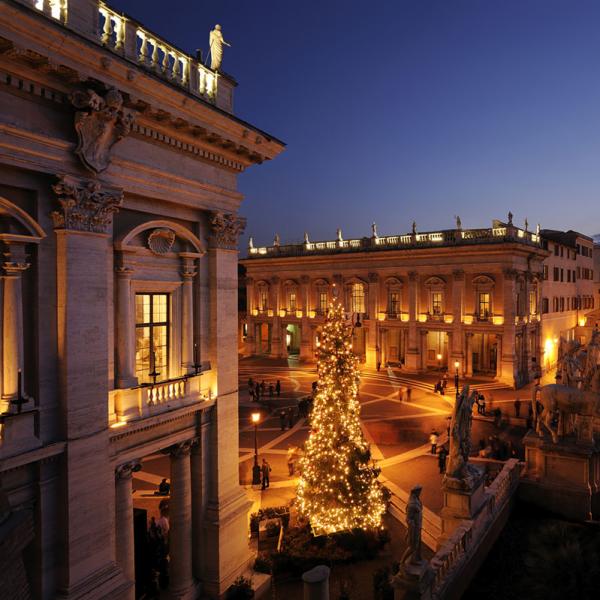 Piazza illuminata con albero di Natale e statue al tramonto.