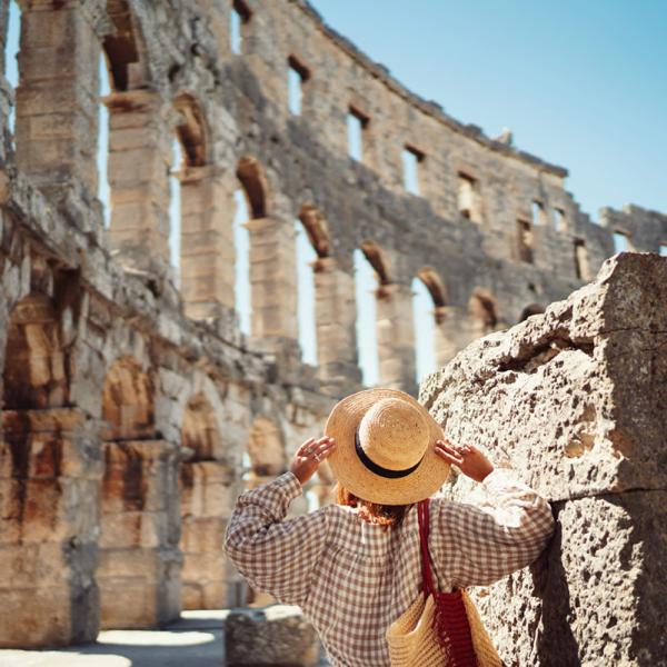 Persona con cappello di paglia ammira antica struttura in pietra sotto il sole.